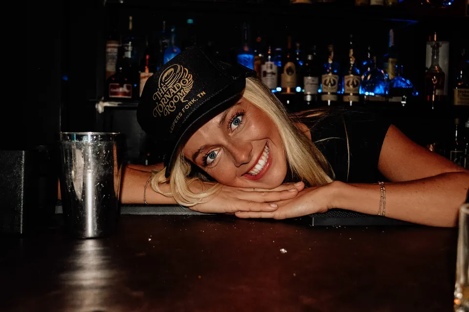 A bartender in a Tornado Room cap leaning on the bar with a grin, the backlit bottles behind.