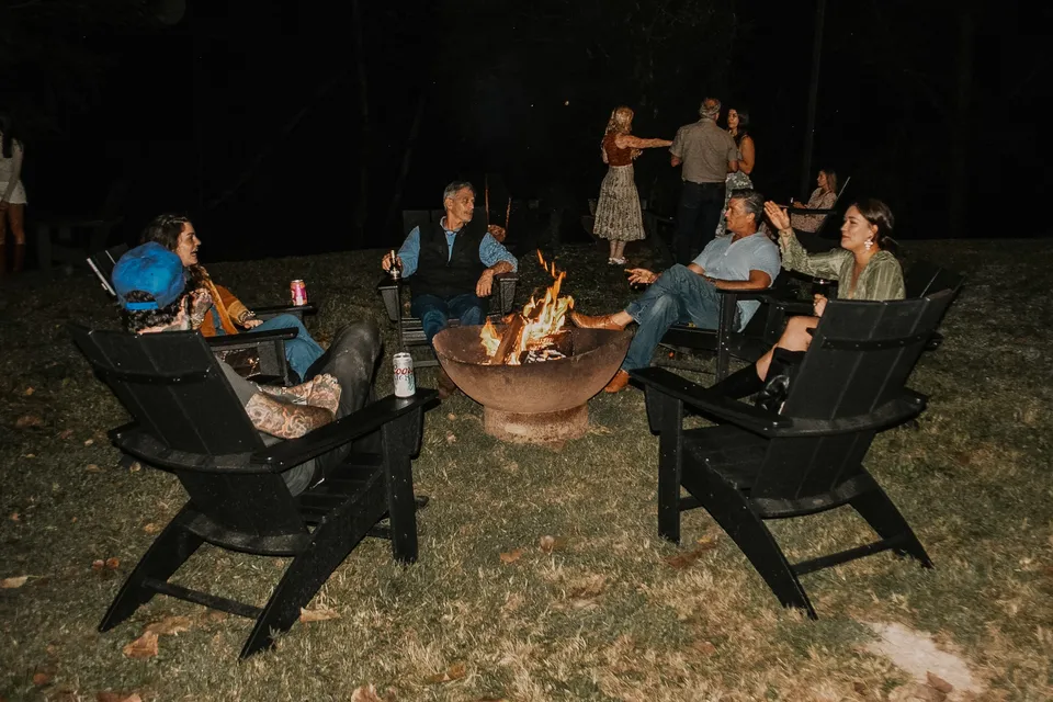 The backyard fire pit after dark: black Adirondack chairs in a ring, drinks in hand.
