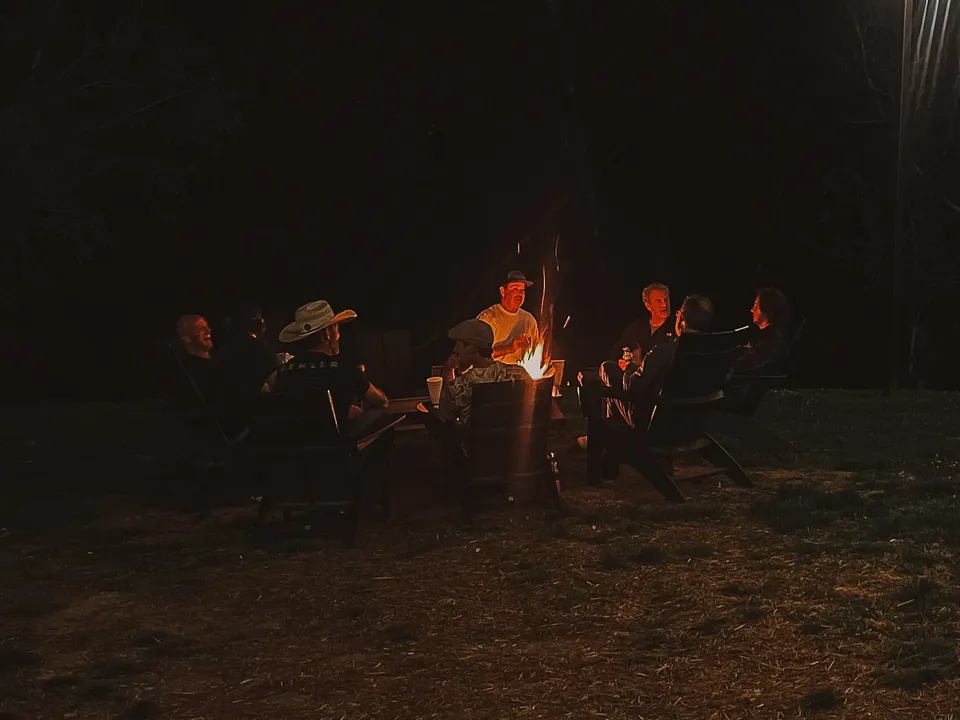 A ring of people in chairs around the fire pit at night, faces lit orange.