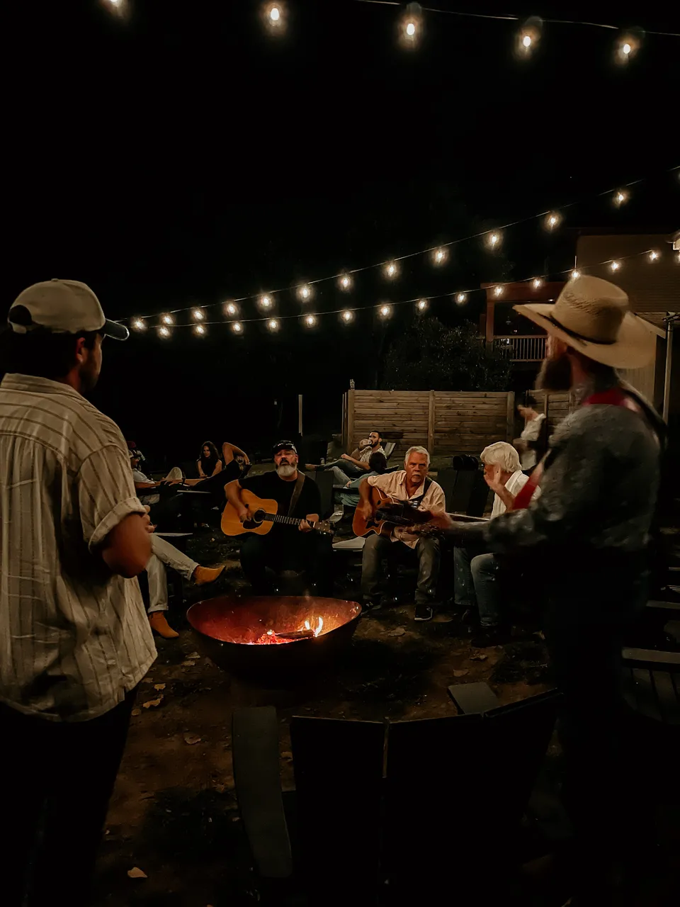 Two players with acoustic guitars in the ring of chairs around the fire pit, string lights overhead, a listener in a cowboy hat standing by.