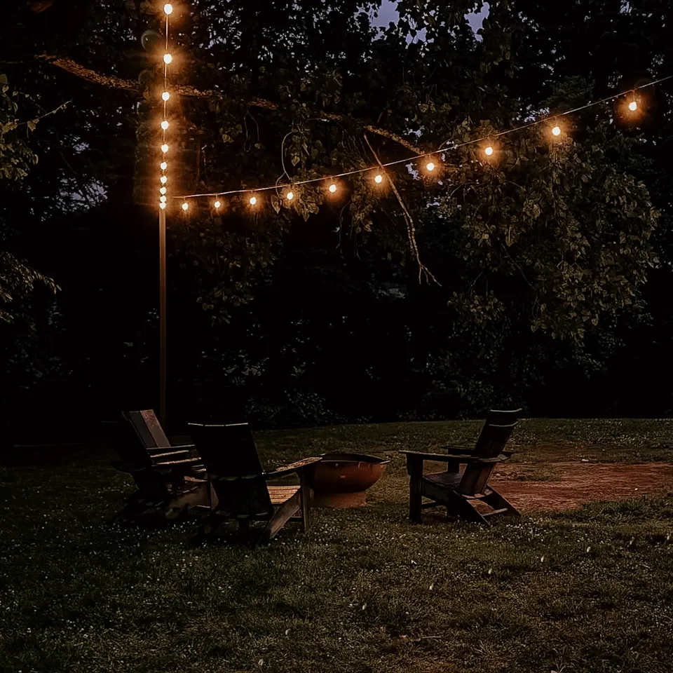 String lights strung from a pole over the empty fire pit and chairs, the trees dark behind.
