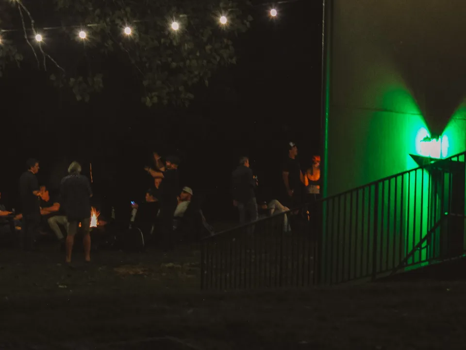 The green lamp’s glow on the corner of the building at night, the stair railing below it, the fire pit crowd out in the dark beyond.